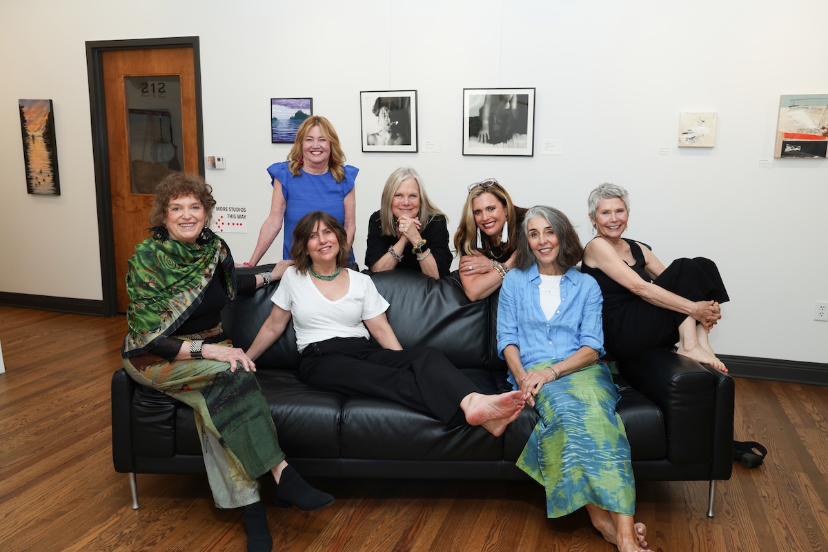Seven women pose together around a black leather couch inside an art gallery with white walls and hardwood floors. Framed artwork is hanging on the wall behind them, and a wooden door with the number "212" is visible in the background.