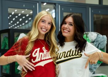 Two young women are smiling side-by-side in an indoor setting. The blonde woman on the left wears a red t-shirt with "Indiana" in white script. The brunette woman on the right wears a white t-shirt with "Purdue" in gold and black script. They are both pointing at their respective shirts. In the background, grey lockers and green and white decorations are visible.