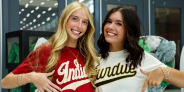 Two young women are smiling side-by-side in an indoor setting. The blonde woman on the left wears a red t-shirt with "Indiana" in white script. The brunette woman on the right wears a white t-shirt with "Purdue" in gold and black script. They are both pointing at their respective shirts. In the background, grey lockers and green and white decorations are visible.