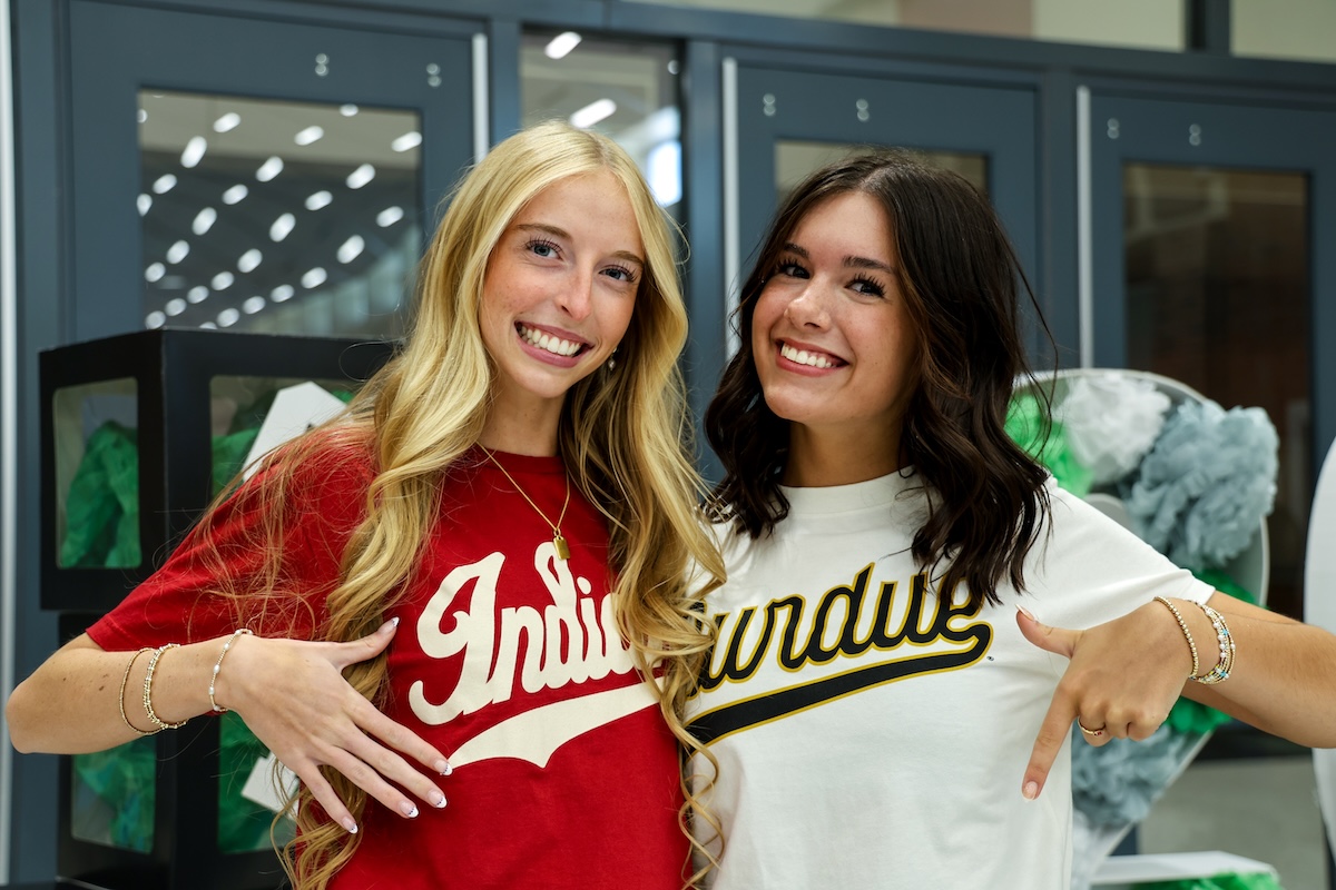 Two young women are smiling side-by-side in an indoor setting. The blonde woman on the left wears a red t-shirt with "Indiana" in white script. The brunette woman on the right wears a white t-shirt with "Purdue" in gold and black script. They are both pointing at their respective shirts. In the background, grey lockers and green and white decorations are visible.