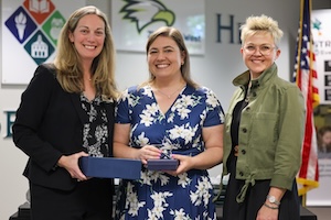 Three women are smiling together at an award presentation. The woman on the left wears a black blazer over a floral top and holds a small blue box. The woman in the center wears a blue and white floral dress and holds a crystal award. The woman on the right wears an olive green jacket over a black top. In the background, there is an American flag and several logos, including an eagle.