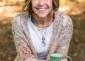 A medium-shot portrait shows a woman with a warm, bright smile sitting outdoors in a wooded area. She has shoulder-length, wavy blonde hair with highlights and is wearing a long, chunky-knit cardigan over a white t-shirt and blue jeans. She is holding a rustic, light green ceramic mug with both hands. Her outfit is completed with layered beaded necklaces, including one with a large black and white oval pendant. The background is softly blurred, showing sun-dappled autumn leaves on the ground.