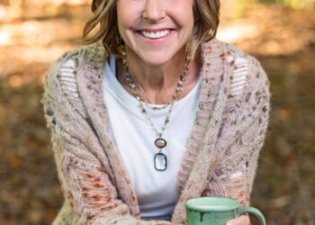 A medium-shot portrait shows a woman with a warm, bright smile sitting outdoors in a wooded area. She has shoulder-length, wavy blonde hair with highlights and is wearing a long, chunky-knit cardigan over a white t-shirt and blue jeans. She is holding a rustic, light green ceramic mug with both hands. Her outfit is completed with layered beaded necklaces, including one with a large black and white oval pendant. The background is softly blurred, showing sun-dappled autumn leaves on the ground.