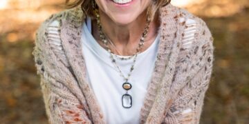 A medium-shot portrait shows a woman with a warm, bright smile sitting outdoors in a wooded area. She has shoulder-length, wavy blonde hair with highlights and is wearing a long, chunky-knit cardigan over a white t-shirt and blue jeans. She is holding a rustic, light green ceramic mug with both hands. Her outfit is completed with layered beaded necklaces, including one with a large black and white oval pendant. The background is softly blurred, showing sun-dappled autumn leaves on the ground.