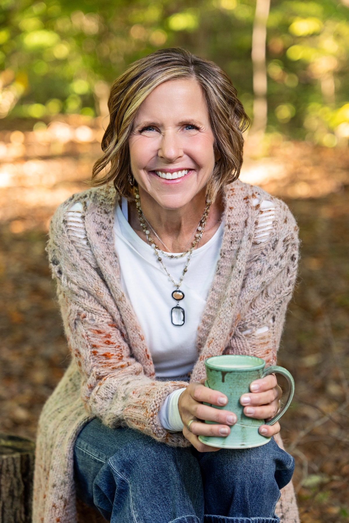 A medium-shot portrait shows a woman with a warm, bright smile sitting outdoors in a wooded area. She has shoulder-length, wavy blonde hair with highlights and is wearing a long, chunky-knit cardigan over a white t-shirt and blue jeans. She is holding a rustic, light green ceramic mug with both hands. Her outfit is completed with layered beaded necklaces, including one with a large black and white oval pendant. The background is softly blurred, showing sun-dappled autumn leaves on the ground.