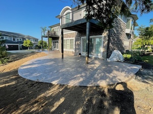 A wide-angle, eye-level shot shows a newly installed stamped concrete patio behind a large, two-story stone and brick home. The tan, curved patio sits beneath a dark wooden deck supported by black pillars. The surrounding yard is currently bare dirt, indicating a project in progress. To the left, a dirt path leads toward the side of the house under a clear, bright blue sky.