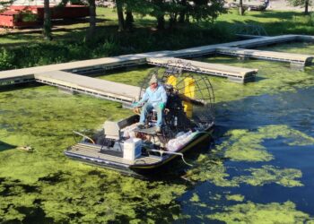 A man in a light blue long-sleeve shirt and a hat operates a flat-bottomed airboat on a waterway covered in thick green algae and duckweed. The boat is equipped with a large rear propeller in a safety cage and several white jugs, possibly containing herbicide or water treatments. A series of floating wooden docks and a grassy bank with trees are visible in the background.
