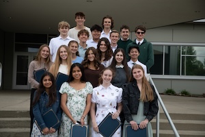 A large group of approximately seventeen smiling high school students stands on concrete steps in front of a modern school building. Several students in the front rows are holding dark blue folders or certificates. They are dressed in a mix of semi-formal and casual attire, including dresses, blouses, and button-down shirts. Large glass windows and a school entrance are visible in the background.