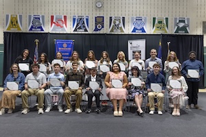 A group of twenty smiling high school students posing with award certificates on a stage. Nine students are seated in the front row, and eleven stand in the back. They are in a gymnasium with black curtains behind them and a row of school sports banners hanging along the top wall. A blue banner for the Rotary Club and an American flag are also visible.