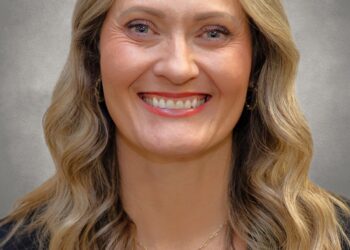 A professional headshot of a woman with long, wavy blonde hair and a bright smile. She is wearing a black blazer over a white top and a thin gold chain necklace. The portrait is set against a neutral, mottled grey background.