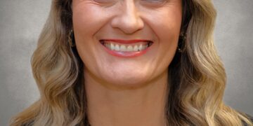 A professional headshot of a woman with long, wavy blonde hair and a bright smile. She is wearing a black blazer over a white top and a thin gold chain necklace. The portrait is set against a neutral, mottled grey background.