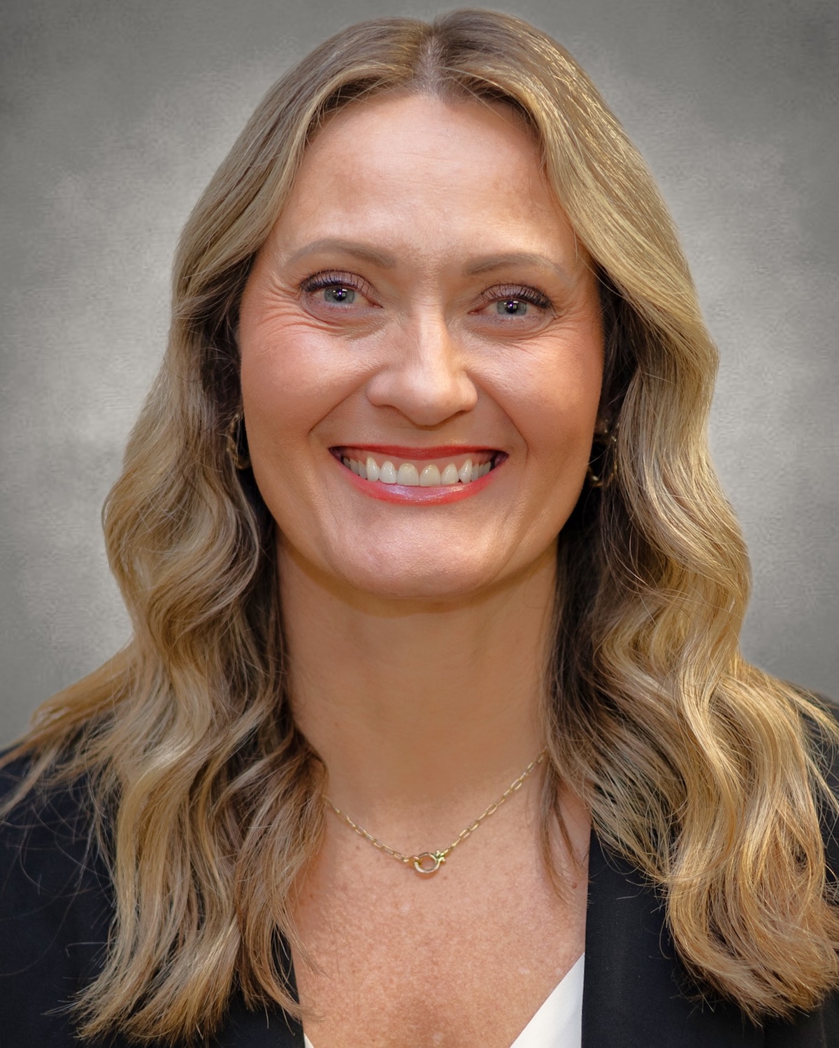 A professional headshot of a woman with long, wavy blonde hair and a bright smile. She is wearing a black blazer over a white top and a thin gold chain necklace. The portrait is set against a neutral, mottled grey background.