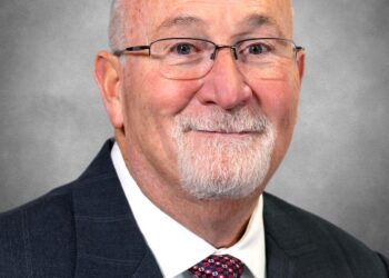 A formal headshot of a man with a white goatee and glasses, smiling warmly. He is wearing a dark pinstripe suit jacket, a white dress shirt, and a red patterned tie. The background is a solid, neutral grey.