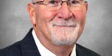 A formal headshot of a man with a white goatee and glasses, smiling warmly. He is wearing a dark pinstripe suit jacket, a white dress shirt, and a red patterned tie. The background is a solid, neutral grey.
