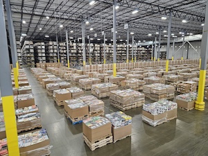 Thousands of books are stacked in large cardboard bins inside a massive, high-ceilinged warehouse. The foreground shows stacks of various hardcovers and paperbacks, including some cookbooks and novels, with a shallow depth of field that blurs the rows of books and yellow support pillars in the background. Overhead, bright industrial lights illuminate the space.