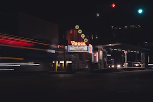 A long-exposure night photograph captures the glowing marquee of the Vogue Theatre in Indianapolis. The classic sign features vertical yellow neon circles above the word "Vogue" in a flowing red script. Below, a bright white marquee board displays text under a dark night sky. To the left, vibrant red and white light trails from passing cars blur across the frame, creating a sense of motion. A green traffic light and a gas station canopy are visible to the right, adding to the urban evening scene.