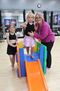 A woman and two young girls pose around a colorful plastic toy slide inside a dance studio. The woman, wearing glasses and a pink long-sleeved shirt, smiles while standing next to a toddler in a black leotard and pink patterned skirt who is standing on top of the slide. To the left, a slightly older girl in a black two-piece dance outfit leans against the slide’s railing. The background shows a mirrored wall, light wood floors, and dance competition posters.