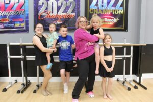 A woman and five children pose in a dance studio in front of a ballet barre. The woman, in a pink long-sleeved shirt and black leggings, holds a young child in her arms. To her left, an older girl holds a toddler, while a young boy in a blue "Nexstar 2025" t-shirt stands between them. To the woman's right stands a young girl in a black dance outfit. The studio has light wood floors and grey walls adorned with large "2022 Champion" and "National Champion" banners from various dance competitions.