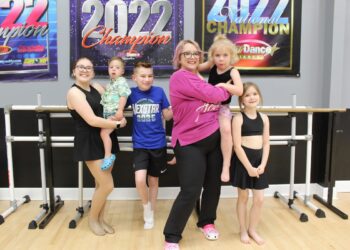 A woman and five children pose in a dance studio in front of a ballet barre. The woman, in a pink long-sleeved shirt and black leggings, holds a young child in her arms. To her left, an older girl holds a toddler, while a young boy in a blue "Nexstar 2025" t-shirt stands between them. To the woman's right stands a young girl in a black dance outfit. The studio has light wood floors and grey walls adorned with large "2022 Champion" and "National Champion" banners from various dance competitions.
