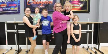 A woman and five children pose in a dance studio in front of a ballet barre. The woman, in a pink long-sleeved shirt and black leggings, holds a young child in her arms. To her left, an older girl holds a toddler, while a young boy in a blue "Nexstar 2025" t-shirt stands between them. To the woman's right stands a young girl in a black dance outfit. The studio has light wood floors and grey walls adorned with large "2022 Champion" and "National Champion" banners from various dance competitions.