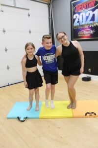 Three young dancers—two girls and one boy—stand with their arms around each other on a colorful foam tumbling mat in a dance studio. The girl on the left and the older girl on the right wear black dance outfits, while the boy in the center wears a blue "Nexstar 2025" t-shirt and black shorts. They are all smiling warmly at the camera. In the background, a white garage-style door and a "2021 Champion" banner are visible against a grey wall.