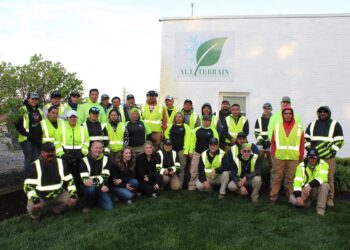 An outdoor group portrait shows about 25 employees of "All Terrain Landscape and Snow Management" posing in front of a white brick building. The workers, a mix of men and women, are dressed in high-visibility neon yellow and green safety vests over dark hoodies or shirts. Some wear baseball caps, and many are smiling. They are arranged in three rows: some kneeling or sitting on the grass in the front, and two rows standing behind them. On the building's wall, a large white sign features the company logo—a green leaf overlapping a light blue snowflake—with the name "ALL TERRAIN" in bold black letters. The scene is softly lit, likely in the early morning or late afternoon, with lush green grass in the foreground and leafy bushes to the left.