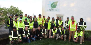 An outdoor group portrait shows about 25 employees of "All Terrain Landscape and Snow Management" posing in front of a white brick building. The workers, a mix of men and women, are dressed in high-visibility neon yellow and green safety vests over dark hoodies or shirts. Some wear baseball caps, and many are smiling. They are arranged in three rows: some kneeling or sitting on the grass in the front, and two rows standing behind them. On the building's wall, a large white sign features the company logo—a green leaf overlapping a light blue snowflake—with the name "ALL TERRAIN" in bold black letters. The scene is softly lit, likely in the early morning or late afternoon, with lush green grass in the foreground and leafy bushes to the left.