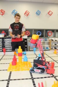 A young man in a "PYRE Robotics" jersey stands behind a competition table, focusing as he operates a handheld controller. In the foreground, a red and black robot with the number "46168A" uses a mechanical arm to lift a yellow and red game piece. The practice field is filled with similar colorful plastic components. The background wall is decorated with red and blue gears and wireframe cubes, alongside organized storage bins for parts.
