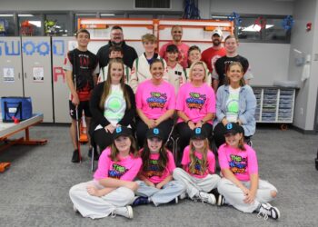 A large group of about 15 students and mentors poses in a modern robotics lab or STEM classroom. In the front row, four young girls sit on the floor wearing neon pink "The Gadget Girlz" t-shirts and black baseball caps with a blue "G" logo. Behind them, four women sit in chairs, two of whom also wear the pink team shirts. The back two rows consist of boys in various team jerseys—including some that say "PYRE Robotics"—and adult male mentors. The background features whiteboards with "TWO" and gear graphics, storage cabinets, and a robotics practice table to the left.