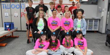 A large group of about 15 students and mentors poses in a modern robotics lab or STEM classroom. In the front row, four young girls sit on the floor wearing neon pink "The Gadget Girlz" t-shirts and black baseball caps with a blue "G" logo. Behind them, four women sit in chairs, two of whom also wear the pink team shirts. The back two rows consist of boys in various team jerseys—including some that say "PYRE Robotics"—and adult male mentors. The background features whiteboards with "TWO" and gear graphics, storage cabinets, and a robotics practice table to the left.