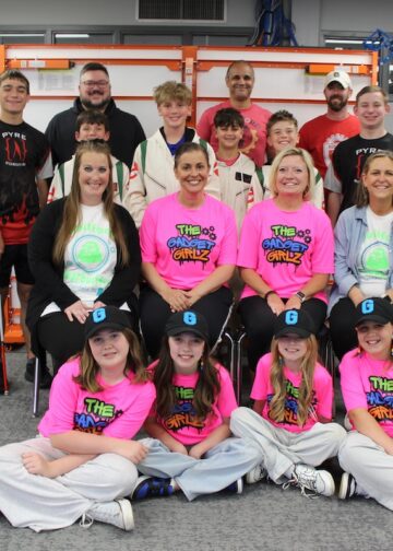 A large group of about 15 students and mentors poses in a modern robotics lab or STEM classroom. In the front row, four young girls sit on the floor wearing neon pink "The Gadget Girlz" t-shirts and black baseball caps with a blue "G" logo. Behind them, four women sit in chairs, two of whom also wear the pink team shirts. The back two rows consist of boys in various team jerseys—including some that say "PYRE Robotics"—and adult male mentors. The background features whiteboards with "TWO" and gear graphics, storage cabinets, and a robotics practice table to the left.