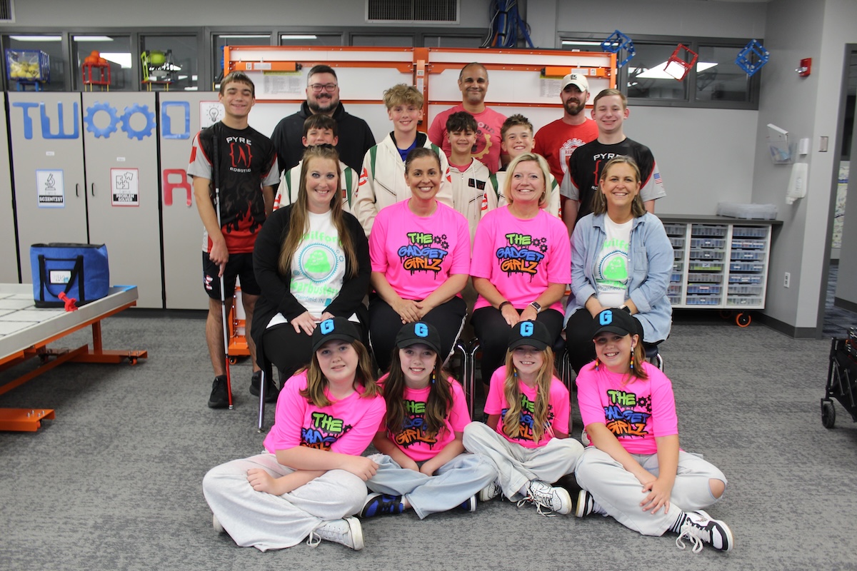 A large group of about 15 students and mentors poses in a modern robotics lab or STEM classroom. In the front row, four young girls sit on the floor wearing neon pink "The Gadget Girlz" t-shirts and black baseball caps with a blue "G" logo. Behind them, four women sit in chairs, two of whom also wear the pink team shirts. The back two rows consist of boys in various team jerseys—including some that say "PYRE Robotics"—and adult male mentors. The background features whiteboards with "TWO" and gear graphics, storage cabinets, and a robotics practice table to the left.