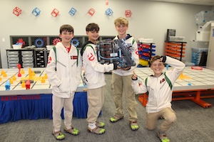 Four young boys in matching white and red team jackets pose in a robotics lab. Three stand in a row, with the two on the right together holding a large, complex robot built from grey and blue components. The fourth boy is kneeling on the right, smiling and gesturing toward the robot. They are positioned in front of a robotics practice table filled with orange and yellow game pieces. The background shows organized storage bins and colorful decorations on the wall.