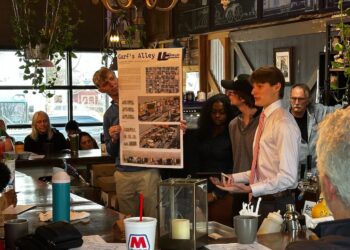 Inside a dimly lit, rustic bar setting, a group of young adults present a design concept to an audience. A young man on the left holds a large poster board titled "Garf's Alley" featuring architectural renderings and floor plans. On the right, a young man in a white button-down shirt and pink tie speaks dynamically to the crowd, flanked by two peers. An older man with glasses watches from behind them, and several audience members sit at the bar counter in the foreground and background.