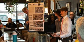 Inside a dimly lit, rustic bar setting, a group of young adults present a design concept to an audience. A young man on the left holds a large poster board titled "Garf's Alley" featuring architectural renderings and floor plans. On the right, a young man in a white button-down shirt and pink tie speaks dynamically to the crowd, flanked by two peers. An older man with glasses watches from behind them, and several audience members sit at the bar counter in the foreground and background.