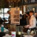 Inside a dimly lit, rustic bar setting, a group of young adults present a design concept to an audience. A young man on the left holds a large poster board titled "Garf's Alley" featuring architectural renderings and floor plans. On the right, a young man in a white button-down shirt and pink tie speaks dynamically to the crowd, flanked by two peers. An older man with glasses watches from behind them, and several audience members sit at the bar counter in the foreground and background.