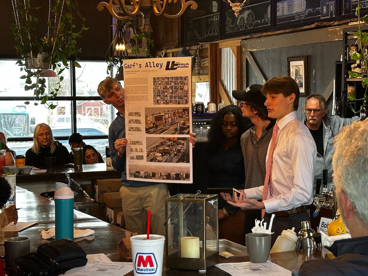 Inside a dimly lit, rustic bar setting, a group of young adults present a design concept to an audience. A young man on the left holds a large poster board titled "Garf's Alley" featuring architectural renderings and floor plans. On the right, a young man in a white button-down shirt and pink tie speaks dynamically to the crowd, flanked by two peers. An older man with glasses watches from behind them, and several audience members sit at the bar counter in the foreground and background.