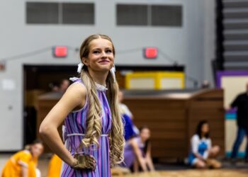 A young woman with long, blonde, curled pigtails tied with white ribbons smiles while posing during a performance. She is wearing a purple and blue striped sleeveless dress with a white ruffled collar and a tan glove on her right hand. The background shows a gymnasium or arena setting with other performers in various colorful costumes slightly out of focus.