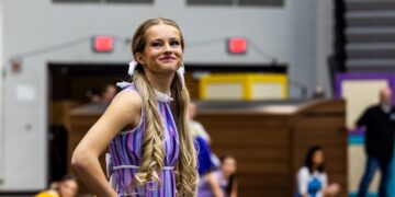 A young woman with long, blonde, curled pigtails tied with white ribbons smiles while posing during a performance. She is wearing a purple and blue striped sleeveless dress with a white ruffled collar and a tan glove on her right hand. The background shows a gymnasium or arena setting with other performers in various colorful costumes slightly out of focus.