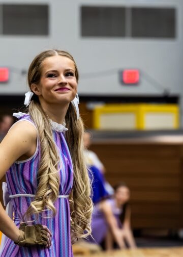 A young woman with long, blonde, curled pigtails tied with white ribbons smiles while posing during a performance. She is wearing a purple and blue striped sleeveless dress with a white ruffled collar and a tan glove on her right hand. The background shows a gymnasium or arena setting with other performers in various colorful costumes slightly out of focus.