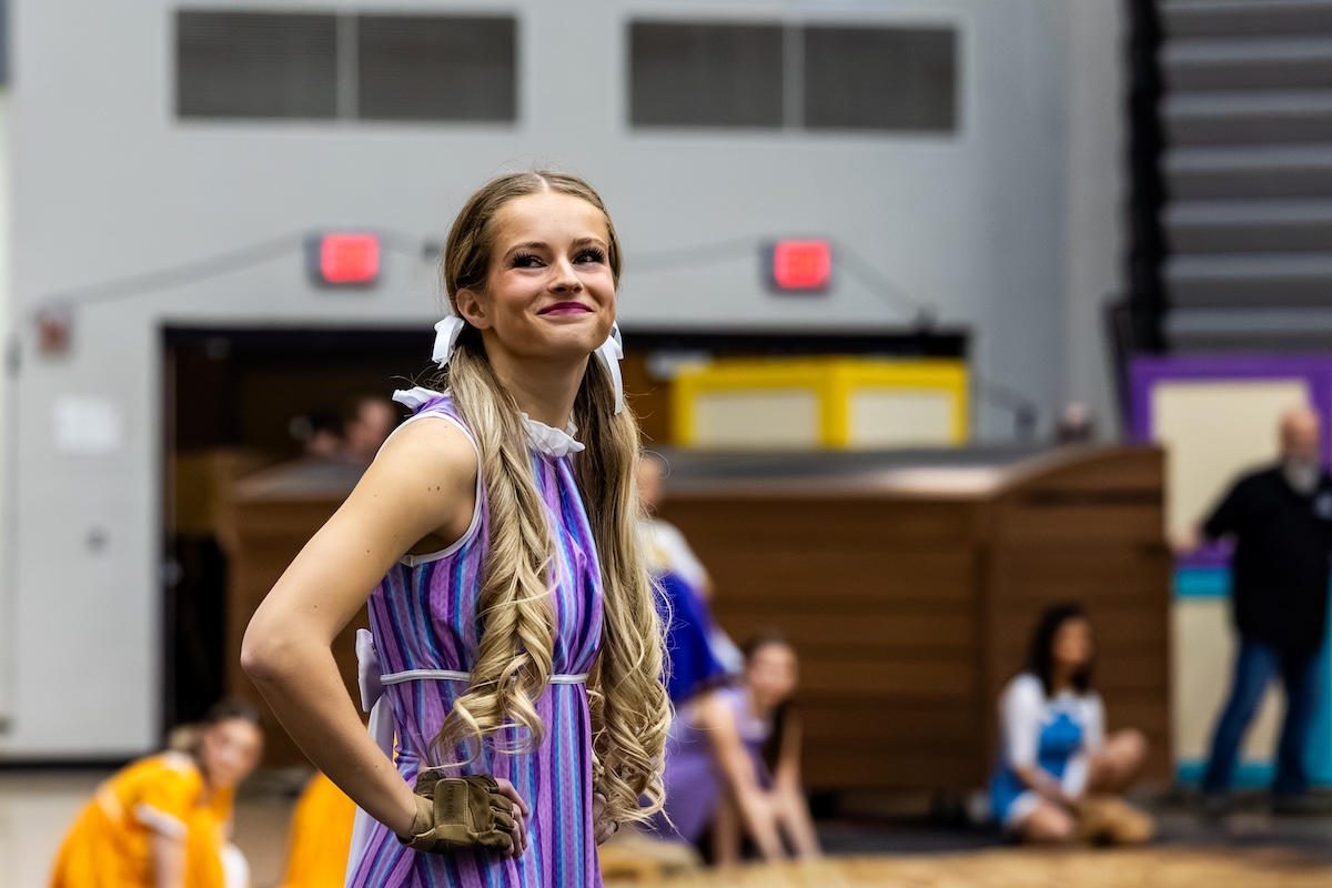 A young woman with long, blonde, curled pigtails tied with white ribbons smiles while posing during a performance. She is wearing a purple and blue striped sleeveless dress with a white ruffled collar and a tan glove on her right hand. The background shows a gymnasium or arena setting with other performers in various colorful costumes slightly out of focus.
