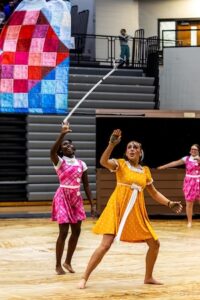 Two color guard performers in vintage-style dresses compete on a gym floor. A girl in a pink plaid dress has just tossed a tall, hooked prop into the air, while a girl in a yellow polka-dot dress tracks its movement with a focused expression. In the background, a large, colorful patchwork quilt-style flag is visible, adding to the theatrical theme.