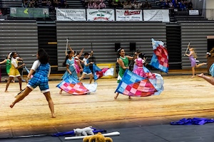 A wide action shot shows a large winter guard ensemble performing a dynamic routine on a gymnasium floor. The performers are dressed in various colorful, vintage-inspired outfits including dresses and rompers in shades of blue, pink, green, and yellow. Many are actively spinning large flags with a vibrant, multi-colored patchwork quilt pattern. The background features bleachers filled with spectators and several large sponsor banners hanging above the performance area.
