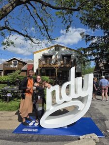 Two people pose with a large, white 3D "ndy" sign (part of a "Love Indy" installation) in a sunny outdoor setting. The man on the left wears an orange shirt and tan pants, while the woman on the right leans against the sculpture, which is mounted on a blue base. In the background, a white two-story building with wooden balconies is visible under a bright blue sky with fluffy white clouds. The area appears to be a lively outdoor patio or courtyard with tables and other people in the distance.