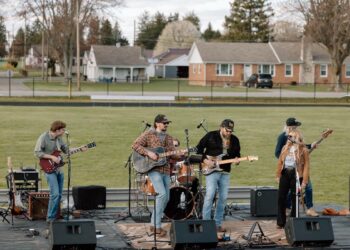 A five-piece live band performs on an outdoor stage set up on an athletic field. The group includes two guitarists, a bassist, a drummer, and a female lead singer. They are dressed in casual attire like flannels, jeans, and hats. In the background, a residential neighborhood with several small houses sits behind a black chain-link fence.