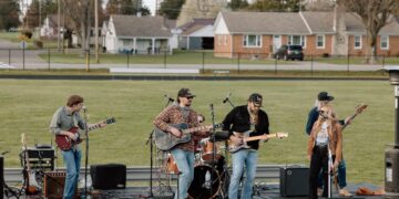 A five-piece live band performs on an outdoor stage set up on an athletic field. The group includes two guitarists, a bassist, a drummer, and a female lead singer. They are dressed in casual attire like flannels, jeans, and hats. In the background, a residential neighborhood with several small houses sits behind a black chain-link fence.