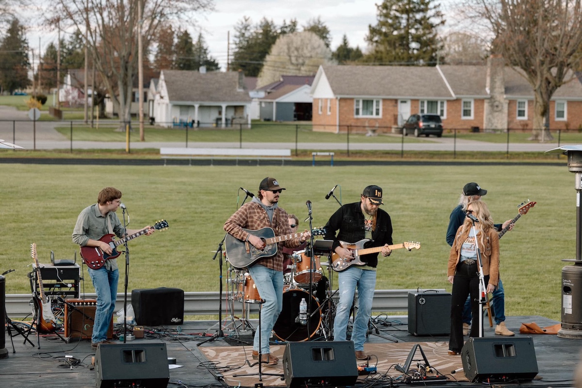 A five-piece live band performs on an outdoor stage set up on an athletic field. The group includes two guitarists, a bassist, a drummer, and a female lead singer. They are dressed in casual attire like flannels, jeans, and hats. In the background, a residential neighborhood with several small houses sits behind a black chain-link fence.