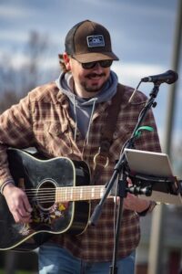 Close-up of a smiling male musician playing an acoustic guitar outdoors. He is wearing a brown trucker hat, sunglasses, and a brown plaid flannel over a grey hoodie. A microphone stand with a mounted tablet for lyrics or music is positioned in front of him.