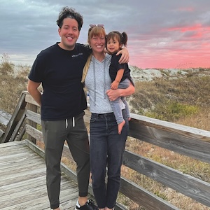 A man, a woman, and a young child pose together smiling on a wooden boardwalk at dusk. The woman holds the child, who wears pigtails, while sand dunes and a vibrant pink-and-grey sunset sky fill the background.