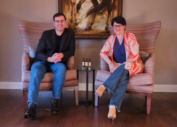 A man in a black blazer and a woman in a patterned orange and white cardigan sit in large patterned wingback chairs. They smile at the camera, with a small round glass table holding three dropper bottles positioned between them.